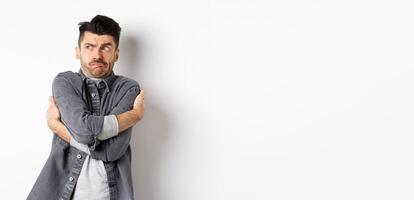 Displeased young man feeling discomfort, hugging himself and staring left at something embarrassing or exposing, standing uncomfortable on white background photo