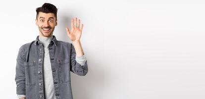 Friendly stylish man saying hello and waiving hand, smiling cheerful, greeting you with hi gesture, standing on white background photo