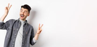Funny young man showing v-signs and smiling at left side, looking at empty space with cheerful face expression, standing on white background photo