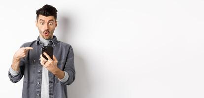 Amazed young man showing cool thing on smartphone, looking and pointing at phone impressed, standing against white background photo