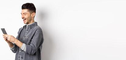 Profile of handsome man chatting on phone, laughing and reading smartphone screen, standing on white background photo