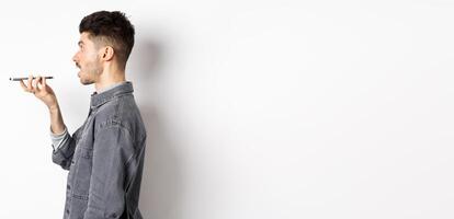 Profile of young man speaking at smartphone, translate with voice-translator app, talking into mobile phone speakerphone, standing on white background photo
