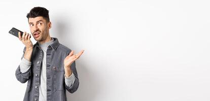 Stylish handsome man talking on speakerphone, record voice message with smartphone near lips, discuss something and looking aside at empty space, standing on white background photo