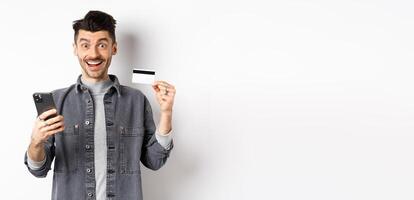 Online shopping concept. Excited man making purchase in internet, easy buying with plastic credit card and smartphone, smiling happy at camera, white background photo