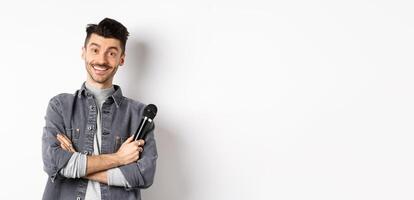 Handsome caucasian guy with moustache cross arms on chest, holding mic and smiling at camera, perform on stage with microphone, standing against white background photo