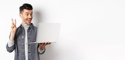 Funny young man chat on laptop, showing v-sign and smiling at computer camera, standing against white background photo