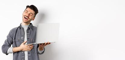 Image of happy guy laughing at funny on laptop, touching belly and smiling satisfied wth good laugh, standing against white background photo