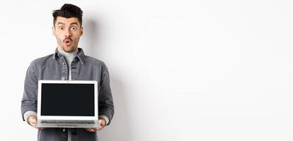 Excited man demostrated empty laptop screen, say wow and look at camera amazed, checking out online offer, showing webpage, standing on white background photo
