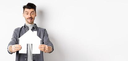 Real estate and insurance concept. Man look with excitement at paper house cutout, buying home, standing on white background photo