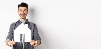 Real estate and insurance concept. Handsome young guy showing paper house cutout and smiling, searching for apartment, buying property, standing against white background photo