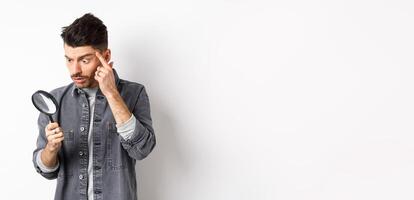 Man looking through magnifying glass and stretching eyelid to see clearly, standing on white background photo