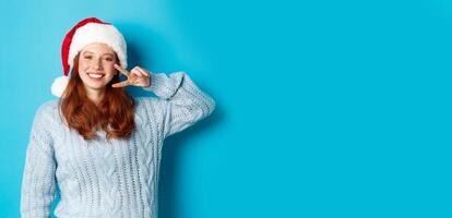 Winter holidays and Christmas Eve concept. Happy teenage girl with red hair, wearing santa hat, enjoying New Year, showing peace sign and smiling, standing over blue background photo