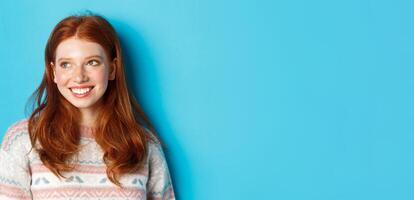 Close-up of beautiful redhead female model in winter sweater, smiling and looking left at copy space, standing over blue background photo