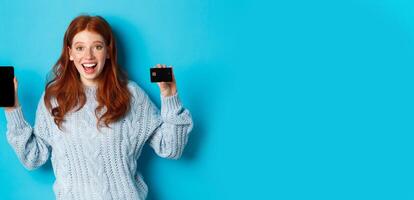 Excited redhead girl showing mobile phone screen and credit card, demonstrating online store or application, standing over blue background photo