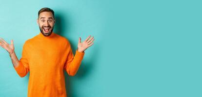 Excited man react to promo offer, spread hands sideways and rejoicing, standing against turquoise background in orange sweater photo