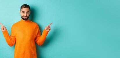 Doubtful and indecisive man pointing fingers sideways, showing different sides, two choices, looking skeptical, standing over light blue background photo