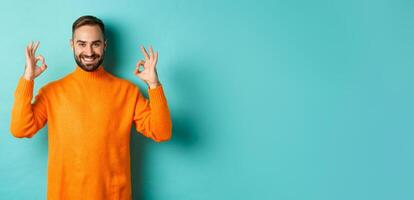 Image of confident smiling man showing okay sign, approve and agree, guarantee quality, standing over light blue background photo