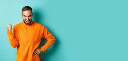 Image of satisfied young man feeling winner, fist pump and saying yes with satisfaction, achieve goal, triumphing while standing over light blue background photo