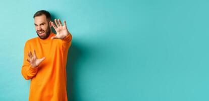 Disgusted man cringe and extending hands in protection, begging for mercy, looking at something with aversion, standing over light blue background photo