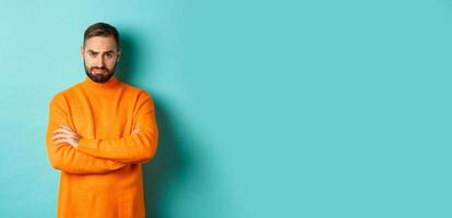 Silly sad guy with beard, sulking and looking offended, cross arms on chest and frowning upset, standing over light blue background photo