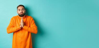 Sulking man begging for help, asking for favour, holding hands in pray and pouting, standing over turquoise background photo
