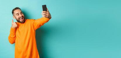 Handsome young man taking selfie on mobile phone, showing thumbs up at smartphone camera, recording vlogg, standing over light blue background photo