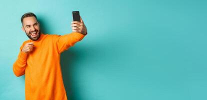 Handsome man calling on mobile phone, taking selfie and pointing at smartphone camera, standing over turquoise background photo