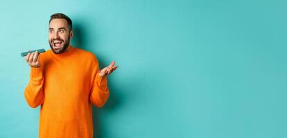 Happy man talking on speakerphone, gesturing and recording voice message on mobile phone, standing in orange sweater over light blue background photo