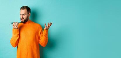 Angry man arguing on speakerphone, record voice message with mad face, standing over light blue background in orange sweater photo