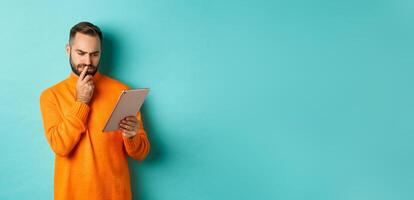 Serious man pondering and looking at digital tablet screen, reading social media, standing over turquoise background photo