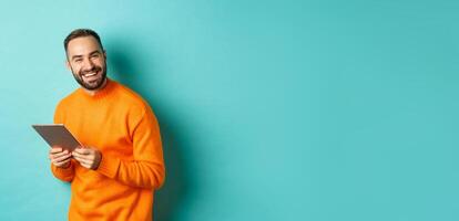 Handsome bearded man using digital tablet, laughing at camera, standing happy against light blue background photo