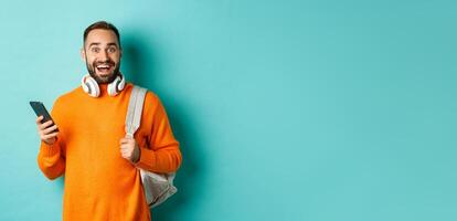 Caucasian man with headphones and backpack staring at camera amazed after reading phone notification, standing over turquoise background photo