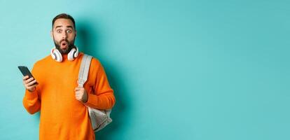 Caucasian man with headphones and backpack staring at camera surprised after reading phone message, standing over turquoise background photo
