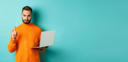 Image of adult man teaching online, showing finger while using laptop, explaining rules, standing over light blue background photo