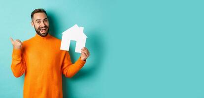 Real estate concept. Satisfied man recommending agency, pointing at copy space banner with thumb, showing paper house maket photo