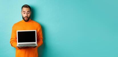 Excited adult male model showing laptop screen, demonstrating online promo, standing happy against light blue background photo