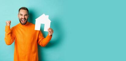 Real estate concept. Excited man saying yes, showing paper house maket and looking satisfied, standing in orange sweater over light blue background photo