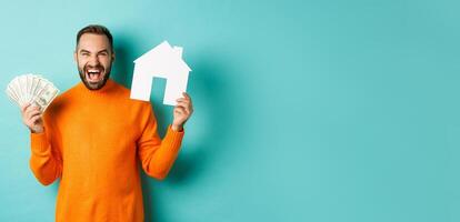 Real estate concept and mortgage concept. Ecstatic caucasian man holding money dollars and house maket, shouting for joy, standing over blue background photo