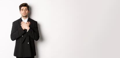 Portrait of dreamy and heartfelt bearded man in suit, close eyes and hold hands on heart, remember something, standing nostalgic over white background photo