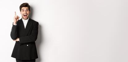 Portrait of attractive man in suit, having an idea, standing excited and raising one finger to tell suggestion, think-up solution, standing over white background photo