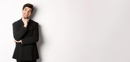 Portrait of thoughtful smiling man in stylish party suit, looking at upper left corner and thinking, imaging things, standing over white background photo