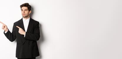 Portrait of serious handsome man in black suit, pointing and looking left at promo banner, showing advertisement on copy space, standing over white background photo