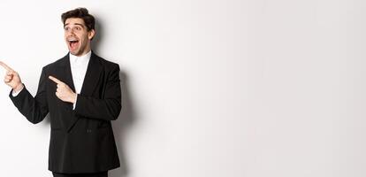 Portrait of amazed and happy handsome man in black suit, pointing and looking left impressed, showing awesome christmas promo, standing over white background photo