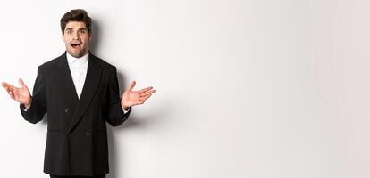 Portrait of confused and worried handsome man in suit, looking at something strange, spread hands sideways and standing puzzled against white background photo