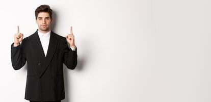 Image of confident and handsome man in formal suit, pointing fingers up, showing copy space on white background photo