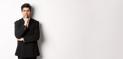Portrait of angry boss in suit shushing at you, telling to be quiet, showing taboo hush sign and frowning, standing over white background photo