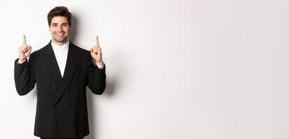 Portrait of attractive caucasian man in stylish black suit, pointing fingers up and smiling, showing christmas advertisement, standing over white background photo