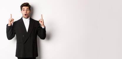 Portrait of surprised handsome businessman in black suit, saying wow and looking amazed, pointing fingers up at copy space, standing over white background photo