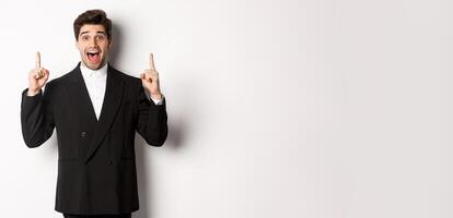 Image of handsome man in party suit, showing holidays promo, pointing fingers up and smiling amazed, standing over white background photo
