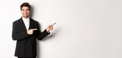 Image of attractive smiling guy dressed for new year party, pointing fingers right and showing advertisement, standing over white background photo